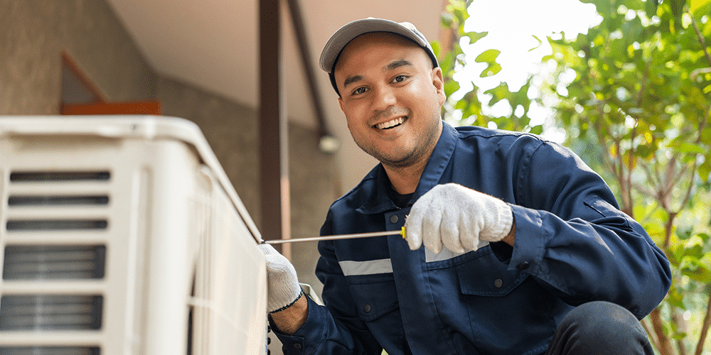 Best HVAC Company Tech in blue shirt and hat smiles as he fixes a residential air conditioner.
