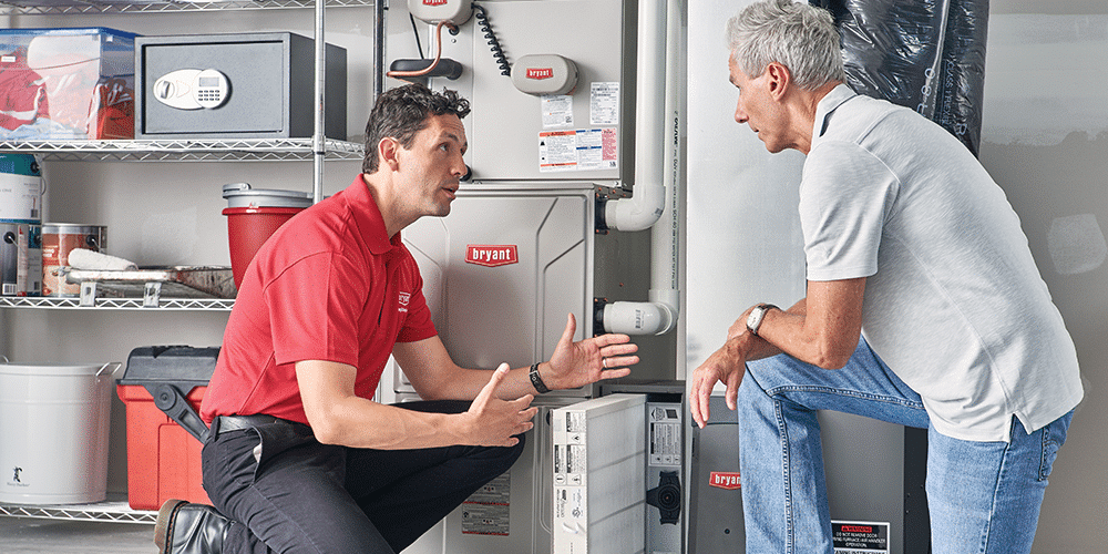 Male technician in red shirt kneels in front of air purifier and explains air quality tips to male homeowner