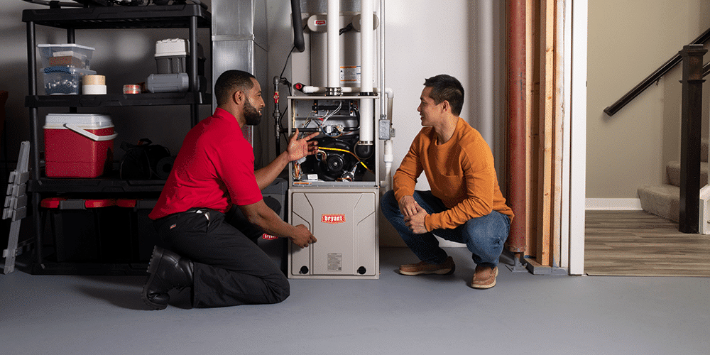 Two men, one a homeowner and one a tech in a red shirt, kneel in front of furnace while tech explains work.