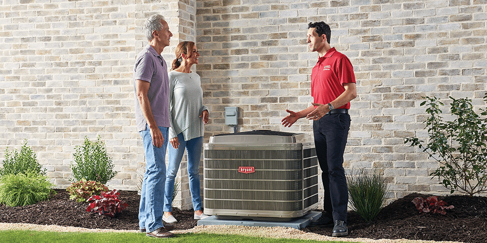 Male technician in red shirt stands with homeowning couple outside and shares air conditioner tips.