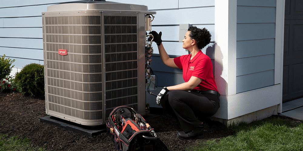Female tech in red shirt performs air conditioner service on residential unit outside house.