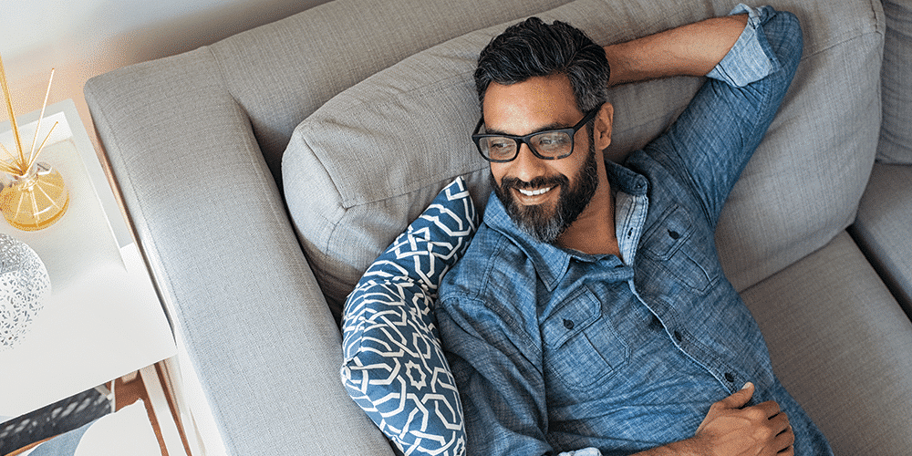 Male homeowner in blue shirt and glasses smiles and lounges on couch in living room.