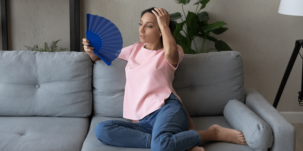 Troubleshoot Uneven Cooling Female homeowner in pink shirt fans herself in living room due to home heating problems.