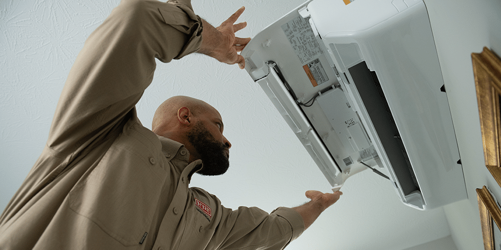 Technician in brown shirt installs ductless HVAC on living room wall.