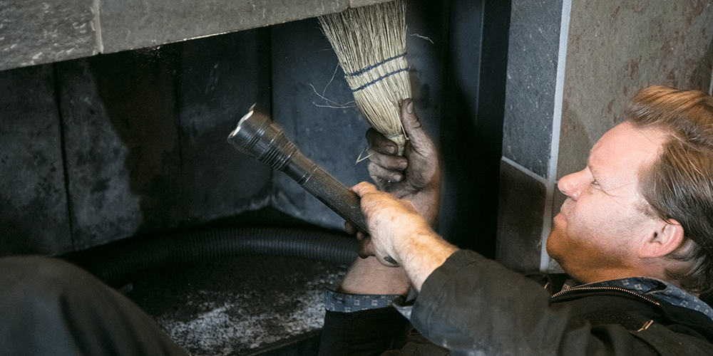 Tech in black jumpsuit performs fireplace maintenance for homeowner.