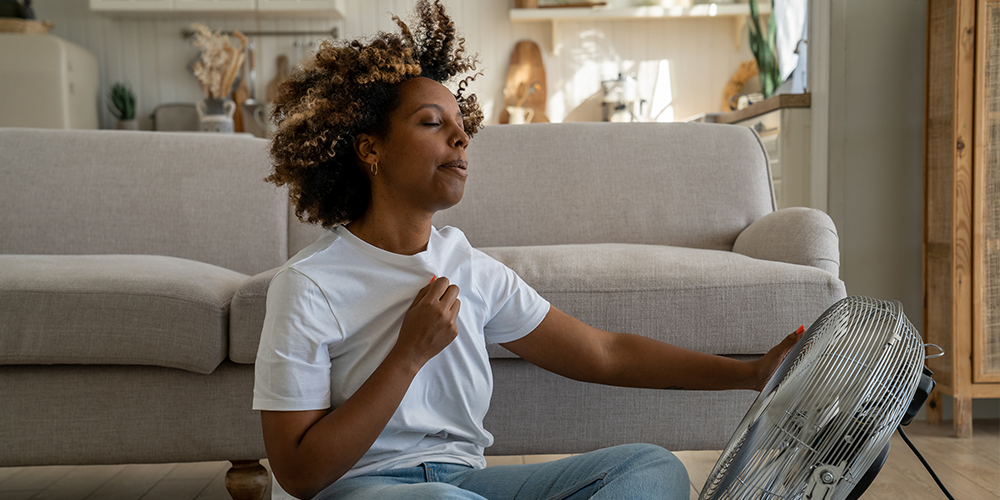 Young homeowning female sits in front of gray couch and uses fan to cool off.