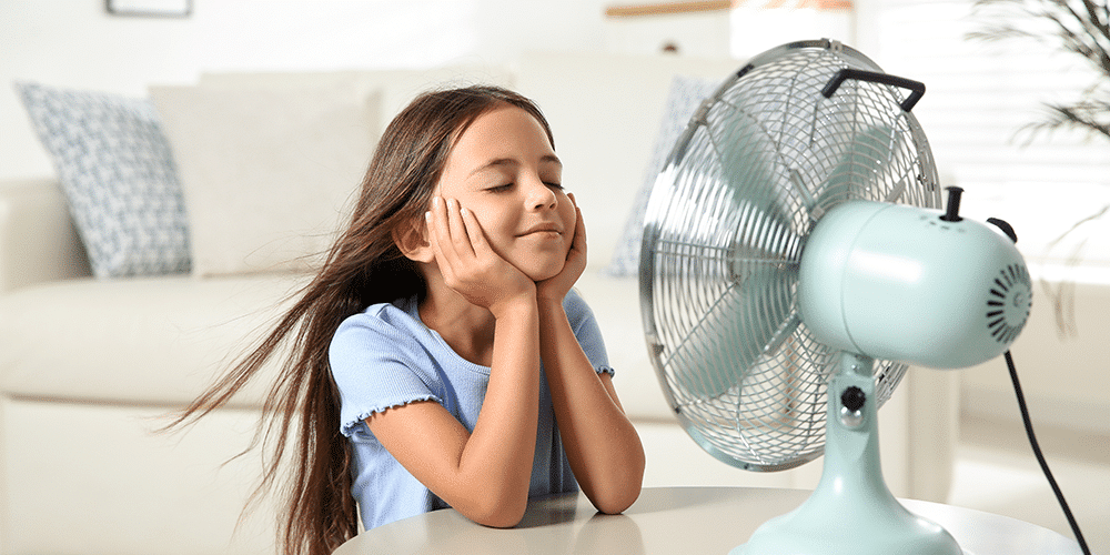 Young girl sits in front of fan in living room to cool off on a hot summer’s day.