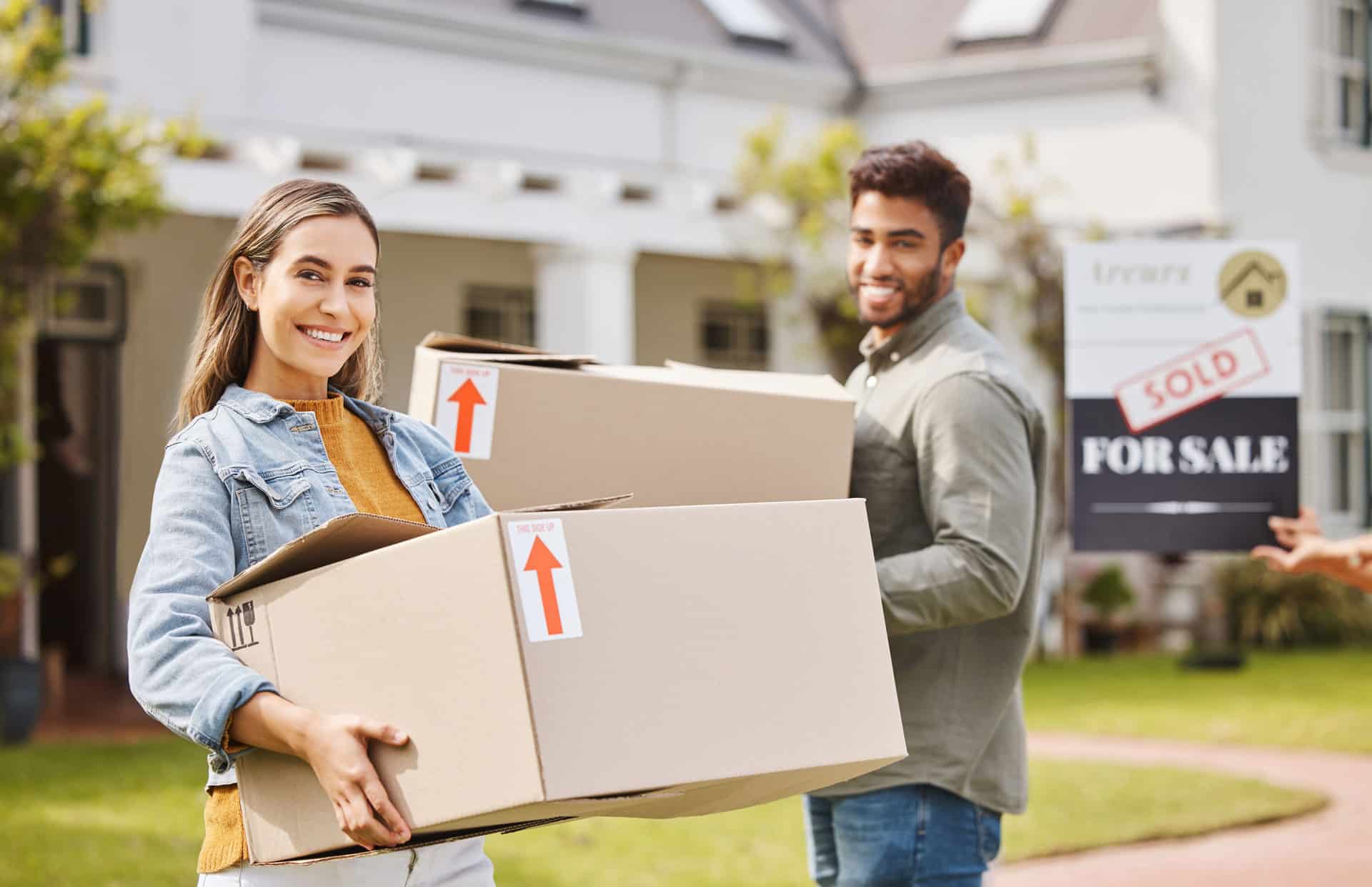 Young homeowning couple moves boxes into their new home with for sale sign out front.