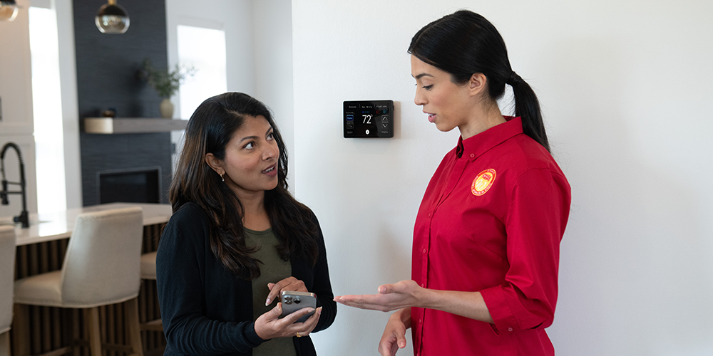Female tech in red shirt walks female homeowner through her new thermostat replacement.