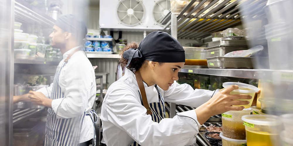 Young female chef stands in commercial fridge looking for food.
