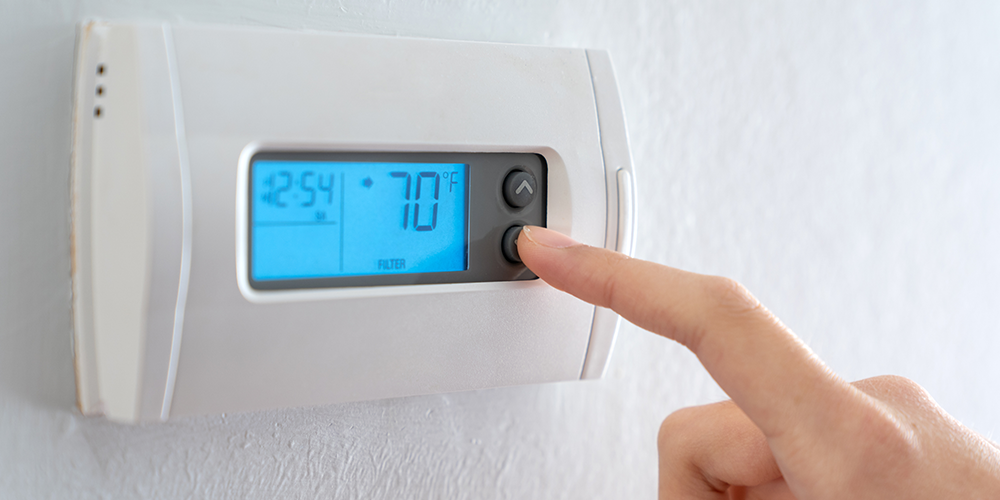 Homeowner's hand and finger adjusts the thermostat settings using the device on their wall.